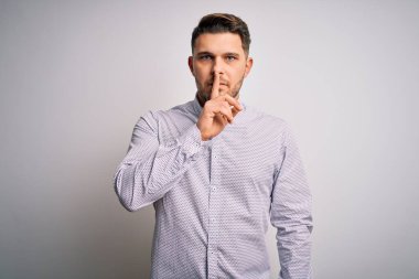 Mavi gözlü genç iş adamı izole edilmiş arka planda duruyor ve parmakları dudaklarında sessiz olmak istiyor. Sessizlik ve gizli konsept.