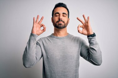 Sakallı, sıradan bir süveter giyen, beyaz arka plan üzerinde duran, gözleri kapalı, parmaklarıyla meditasyon hareketi yapan genç yakışıklı bir adam. Yoga konsepti.