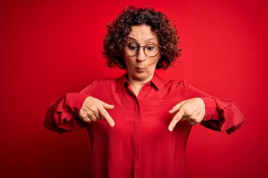 Orta yaşlı, kıvırcık saçlı, rahat gömlekli ve kırmızı arka planda gözlüklü güzel bir kadın. Parmaklarını göstererek reklamını yapıyor, şaşırmış yüzünü ve ağzını açıyor.