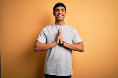 Yakışıklı Afro-Amerikan sporcu sarı arka planda spor giysisi giyerek ellerini kavuşturup af diliyor kendinden emin bir şekilde gülümsüyor..