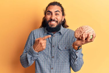 Young arab man holding brain smiling happy pointing with hand and finger  clipart