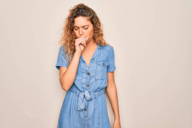 Mavi gözlü genç bir kadın beyaz arka plan üzerine günlük kot elbise giyiyor kendini iyi hissetmiyor ve soğuk algınlığı veya bronşitin belirtisi olarak öksürüyor. Sağlık hizmeti kavramı.