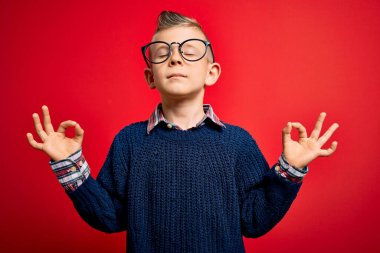 Mavi gözlü, kırmızı arka planda akıllı gözlük takan, gözleri kapalı, meditasyon yaparken parmaklarıyla gülümseyen küçük beyaz bir çocuk. Yoga konsepti.