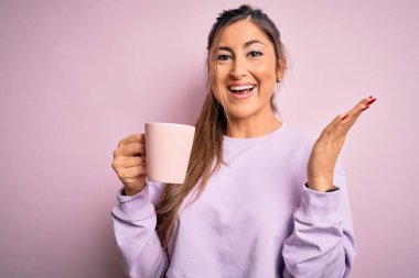 Genç, güzel esmer bir kadın izole edilmiş pembe arka planda bir kupa kahve içiyor çok mutlu ve heyecanlı, kazanan ifade zaferi kutluyor büyük bir gülümsemeyle ve el kaldırarak.