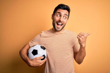 Sakallı yakışıklı oyuncu, sarı arka planda futbol topu tutuyor, gülümsüyor ve başparmağıyla yan tarafı işaret ediyor..