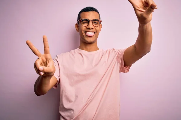 ピンクの背景にカジュアルなTシャツと眼鏡を身に着けているアフリカ系アメリカ人の男性が舌で笑っていて 両手の指が勝利サインをしているのを見せている 第二番 — ストック写真