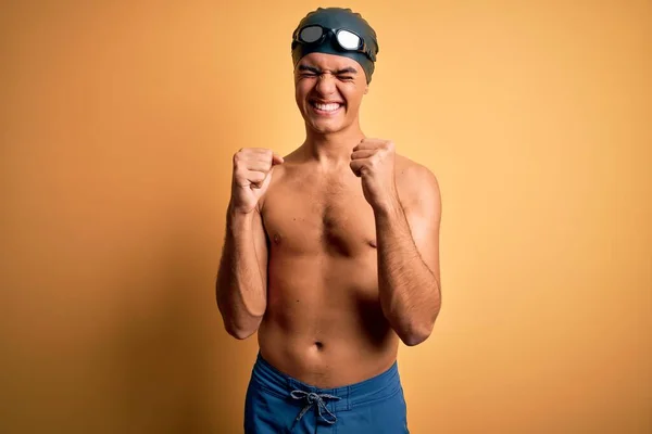 年轻英俊的男子赤身裸体 身穿泳衣 头戴一顶淡黄的泳帽 为成功而兴奋 举起双臂 闭上双眼 微笑着庆祝胜利 赢的概念 — 图库照片