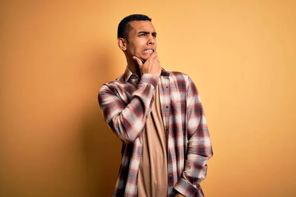 年轻英俊的美国黑人男子穿着休闲衫站在黄色背景下思考着一个问题 手托下巴忧心忡忡 — 图库照片