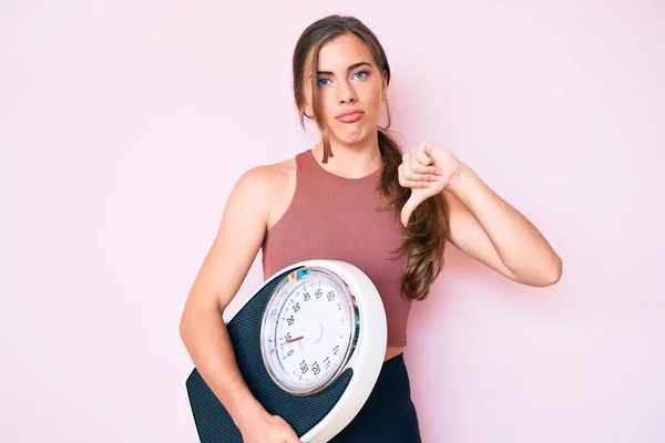 怒った顔で体重減少とバランスをとるために体重計を持っている美しい若い白人女性 親指で嫌いを示す負の記号ダウン 拒絶概念 — ストック写真
