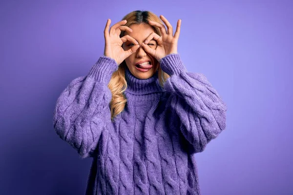 紫の背景にカジュアルなタートルネックセーターを着た若い美しいブロンドの女性は 双眼鏡が舌を突き出すようなOkジェスチャーをして 目は指を通して見ています 狂気の表現 — ストック写真