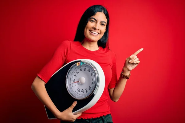 Young beautiful hispanic fitness woman holding scale for healthy weight over red background very happy pointing with hand and finger to the side