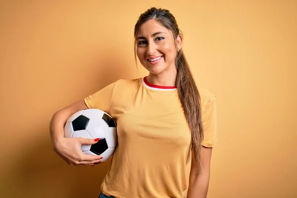 美しいブルネットのプレーヤーの女性プレイサッカーボールを使用して黄色の背景に幸せな顔立ちと笑顔で自信を持って笑顔を示す歯 — ストック写真