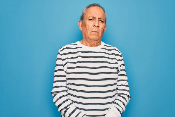 Senior handsome grey-haired man wearing striped sweater over isolated blue background with serious expression on face. Simple and natural looking at the camera.