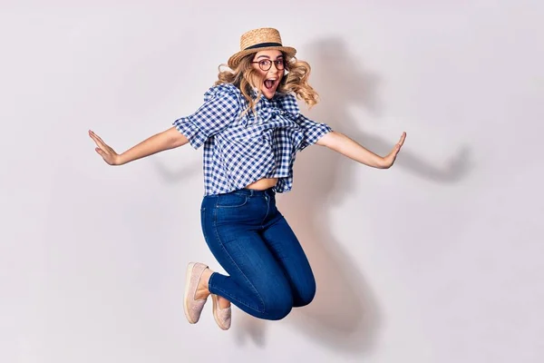 Young beautiful blonde woman wearing glasses and summer hat smiling happy. Jumping with smile on face over isolated white background.