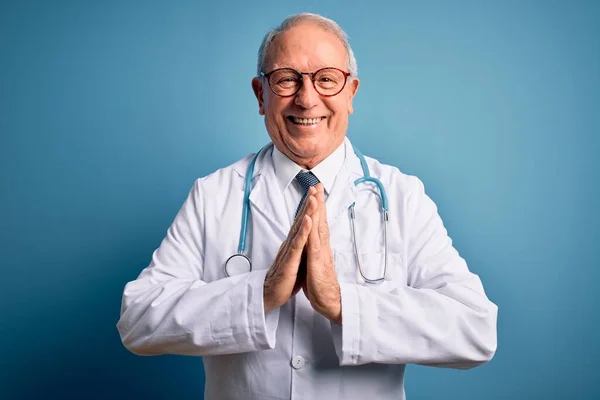 シニア白髪の医師男身に着けています聴診器と医療コート上の青の背景祈る上に手で一緒に求めて許し笑顔自信 — ストック写真
