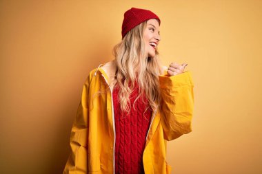 Genç, sarışın, yağmurluk ve yünlü şapka giyen, izole edilmiş sarı arka planda gülümseyen, gülümseyen ve baş parmağıyla yüzünü gösteren güzel bir kadın..