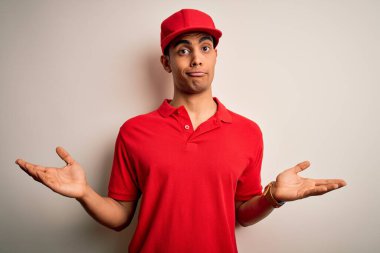 Genç, yakışıklı, Afro-Amerikan bir adam kırmızı arka plan üzerinde sıradan polo ve şapka takıyor ve elleri ve kolları havada, kafası karışmış bir ifade takınıyor. Şüphe kavramı.