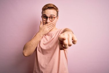 Genç ve yakışıklı kızıl tişörtlü bir adam pembe arka planda dikiliyor sana gülüyor, kamerayı işaret ediyor, ağzının üzerinde utanç ifadesi var.
