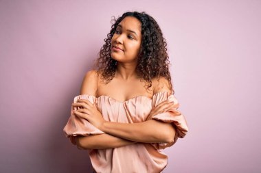 Kıvırcık saçlı, rahat tişörtlü pembe arka planda duran kolları çapraz ve kendinden emin genç bir kadın.
