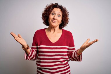 Orta yaşlı güzel kıvırcık saçlı kadın beyaz arka planda sıradan çizgili süveter giyiyor ve elleri ve kolları havada kafası karışmış bir ifade takınıyor. Şüphe kavramı.