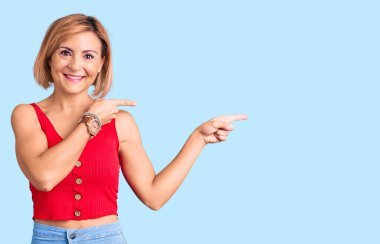 Gündelik kıyafetler giyen genç sarışın kadın gülümseyip kameraya bakıyor ve iki eliyle ve parmaklarıyla kenarı işaret ediyor.. 