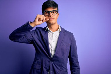 Ceket ve gözlük takan genç yakışıklı iş adamı izole edilmiş mor arka plan ağzı ve parmaklarıyla fermuar gibi kapalı dudaklar. Gizli ve sessiz, tabu konuşması