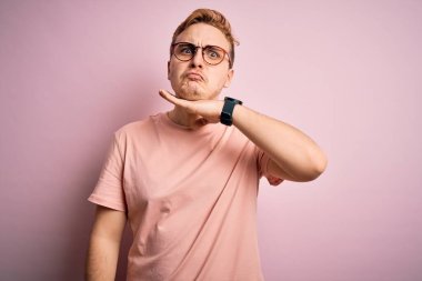 Genç, yakışıklı, günlük tişört giyen, pembe arka planda duran, boğazı bıçak gibi kesen, saldırganlığı şiddetli bir şiddetle tehdit eden bir adam.
