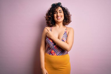 Genç, güzel bir Arap kadın mayo giyiyor ve pembe arka planda güneş gözlüğü takıyor. Gülümsüyor ve gülümsüyor. Yüzünde mutlu ve doğal bir ifade var.