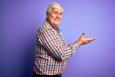 Üst düzey yakışıklı beyaz gömlekli, izole edilmiş mor arka planda, avuç içleri açık, fotokopi alanını gösteren, gülümseyen, heyecanlı mutlu bir gülümseme sunan bir adam.