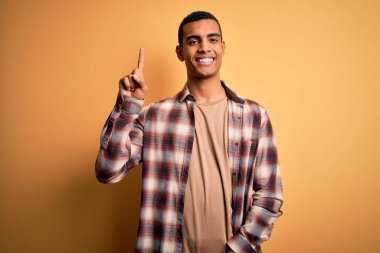 Sarı arka planda dikilen ve başarılı bir fikri işaret eden gündelik gömlekli Afro-Amerikan genç adam. Heyecanlı ve mutlu. Bir numara..