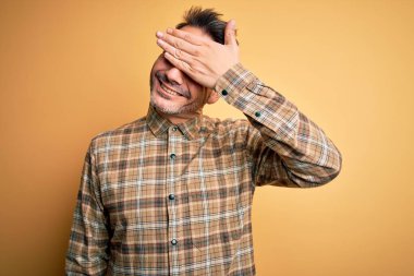 Sarı arka plan üzerinde dikilen rahat tişörtlü genç yakışıklı adam. Gülümsüyor ve elleri yüzünde şaşkınlıktan gözleri kapatıyordu. Kör kavram.