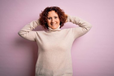 Orta yaşlı güzel kıvırcık saçlı kadın pembe arka planda rahat rahat kazak giyiyor kolları ve elleri başının arkasında mutlu bir şekilde gülümsüyor.