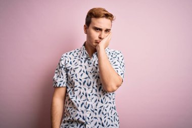 Genç, yakışıklı, yaz gömleği giyen, pembe arka planda duran, kollarını kavuşturarak depresyondan sıkılmış ve yorgun görünen bir adam..