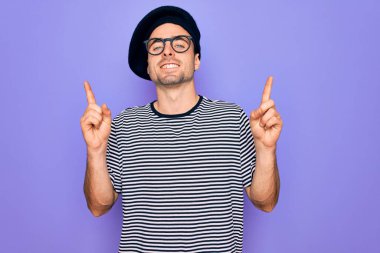 Mor arka planda mavi gözlü, çizgili tişört ve Fransız beresi giyen, kendine güvenen, parmakları farklı yönleri gösteren yakışıklı bir adam. Reklam için boşluk kopyala