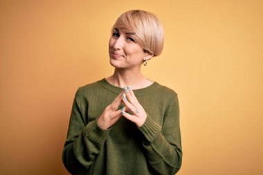 Modern kısa saçlı genç sarışın kadın sarı arka planda sıradan bir süveter giyiyor eller yan yana ve parmaklar çapraz gülümseyerek gevşemiş ve neşeli. Başarı ve iyimser