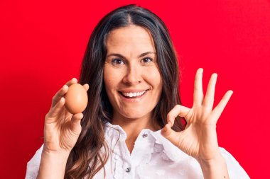 Güzel esmer iş kadını elinde bir deste parayla kırmızı arka planda imza atıyor parmaklarıyla tamam işareti yapıyor, gülümseyerek mükemmel bir sembol oluşturuyor.