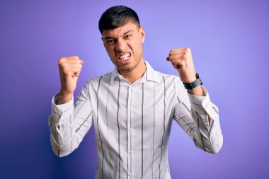 Zarif bir iş gömleği giyen yakışıklı genç adam mor arka planda kızgın ve kızgın duruyor öfkeyle bağırırken kızgın ve öfkeli yumruklar sallıyor. Öfke ve agresif kavram.