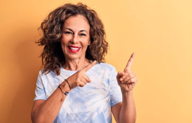 Orta yaşlı güzel bir kadın sarı arka planda dikilen günlük boyalı tişört giyiyor gülümsüyor ve kameraya bakıyor iki eliyle ve parmaklarıyla yan tarafı işaret ediyor..