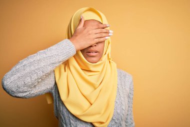 Genç ve güzel Afro-Amerikalı kız, izole edilmiş sarı arka plan üzerine Müslüman başörtüsü takıyor. Gözlerini eliyle kapatıyor, ciddi ve üzgün görünüyor. Görme, saklama ve reddetme konsepti