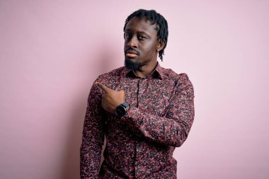 Genç, yakışıklı, Afro-Amerikalı bir adam pembe arka planda duran, ciddi ve sakin bir yüz ifadesi sergileyen, el parmağı ile pembe gömlekli bir adam.