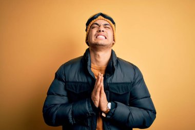 Genç Brezilyalı kayakçı kar sporu kıyafetleri ve sarı arka planda kayak gözlüğü takıyor. Elleri bağlı yalvarıyor ve dua ediyor. Yüzünde umut ifadesi var. Yalvarıyorum..