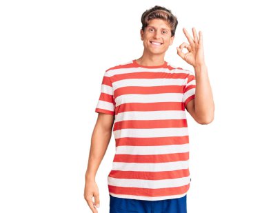 Gündelik kıyafetler giyen, gülümseyen, el ve parmaklarıyla imza atan yakışıklı bir genç. başarılı ifade. 