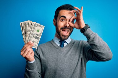 Sakallı genç yakışıklı bir adam mavi arka planda bir deste banknot tutuyor, gülümsüyor ve el pençe divan duruyor.