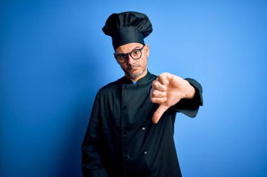 Aşçı üniforması giyen genç ve yakışıklı bir aşçı ve izole edilmiş mavi arka planda şapkası olan mutsuz, kızgın, reddedilmiş ve olumsuz jestler yapan bir adam. Kötü ifade.