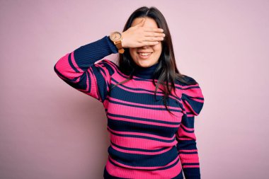 Genç, esmer, zarif bir kadın pembe izole edilmiş arka planda çizgili tişört giyiyor, gülümsüyor ve elleri yüzünde şaşkınlıktan gözleri kapayarak gülüyor. Kör kavram.