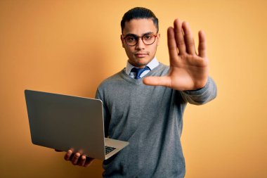 Genç Brezilyalı iş adamı, dizüstü bilgisayarla izole edilmiş sarı arka planda duruyor ve ciddi ve kendinden emin bir ifadeyle dur işareti yapıyor, savunma hareketi yapıyor.