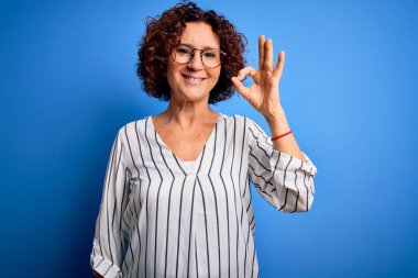 Orta yaşlı güzel kıvırcık saçlı kadın izole edilmiş arka planda sıradan çizgili tişört giyiyor. Gülümsüyor ve el ve parmaklarıyla iyi işareti yapıyor. Başarılı ifade.