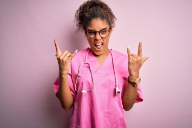 Tıbbi üniforma giyen Afrikalı Amerikalı hemşire kız ve pembe arka planda steteskop takmış çılgın bir ifadeyle elleri havada rock sembolü yaparak bağırıyor. Müzik yıldızı. Ağır konsept.