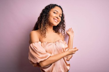 Kıvırcık saçlı, pembe tişörtlü genç ve güzel bir kadın pembe arka planda durup gülümseyerek içeri girmeye davet ediyor.