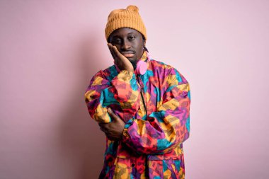 Genç, yakışıklı, Afro-Amerikan bir adam pembe arka plan üzerinde renkli bir ceket ve şapka giyiyor. Kollarını kavuşturup depresyondan sıkılmış ve yorgun görünüyor..